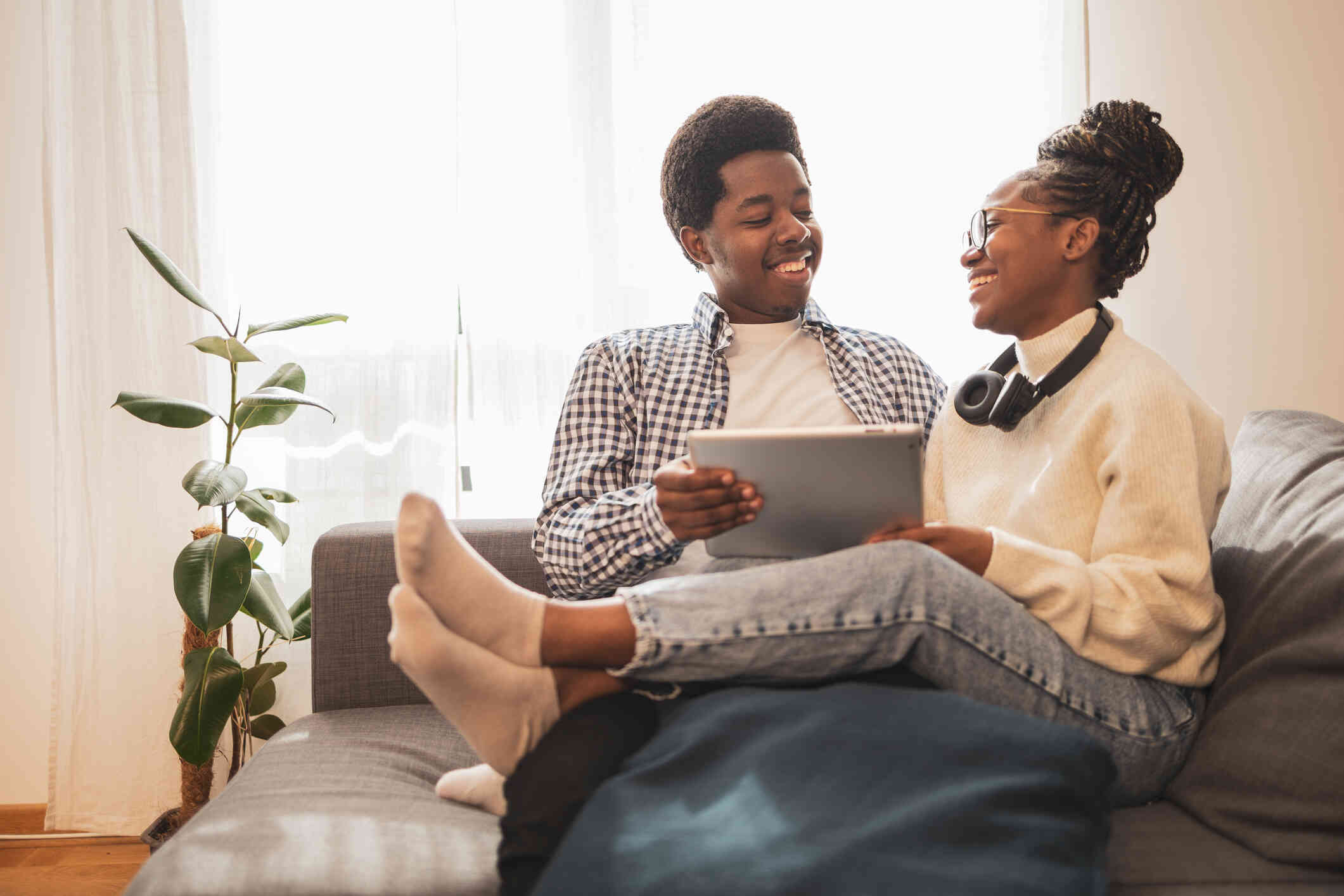 A male and female couple sit on the couch together with the female legs across her partners lap while they hold a tablet and smile at one another.