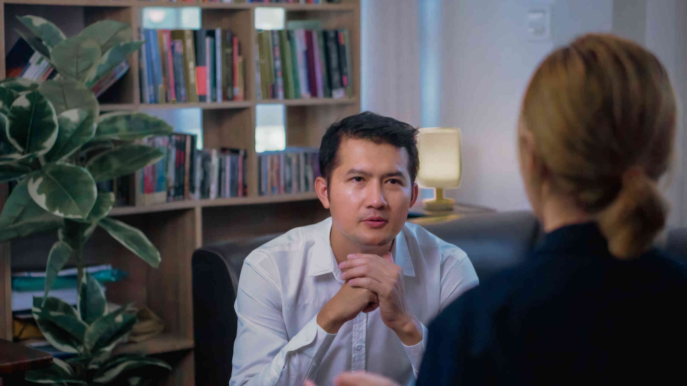 A man in a white button down shirt leans forward while sitting on the couch and claspsp his hands together as he looks at the therapist sitting across from him.