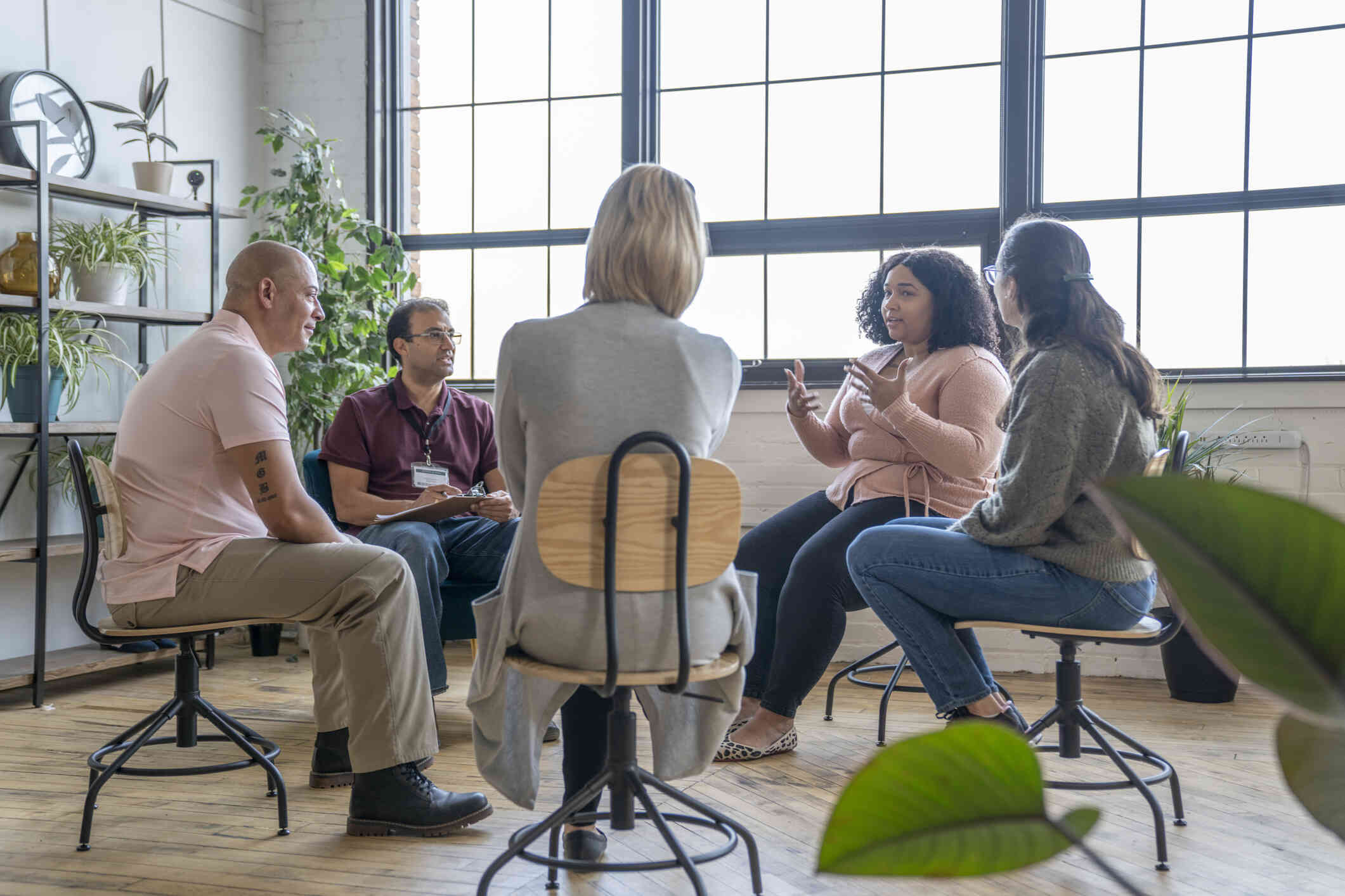 Five adults sit in chairs in a circle as one woman talks during a group therapy session.